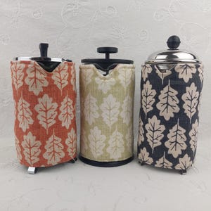 May include: Three French presses with fabric sleeves. The sleeves feature leaf patterns in orange, green, and navy blue. The presses have black handles and silver tops. The presses are on a white surface.