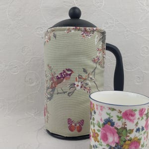 May include: A French press coffee maker with a black handle and lid, and a floral bird design cosy. A matching floral teacup is next to the coffee maker. The cosy has a bird and butterfly design with pink, purple, and green accents.