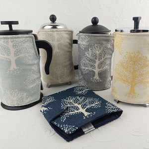 May include: Four French press coffee makers with fabric covers featuring a tree pattern. The covers are in blue, gray, beige, and yellow. A matching fabric coaster is also included.