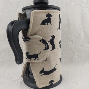 May include: A beige fabric coffee press cosy with a black dachshund silhouette pattern. The cosy has a strap to secure it to the coffee press.