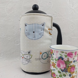 May include: A French press coffee maker with a cream-colored cozy featuring cat faces with glasses and blue striped cat faces. A floral patterned mug is next to the coffee maker. The coffee maker has a black handle and lid.