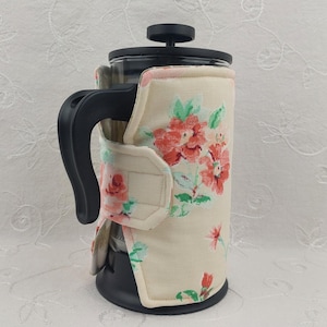 May include: A French press coffee maker with a floral cozy. The cozy is cream-colored with a pattern of red and pink flowers and green leaves. The handle and lid are black.