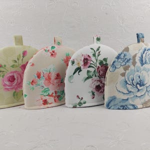 May include: Four floral tea cozy covers in different colors and patterns. The covers are made of fabric with a floral print. The covers are in the colors of green, beige, white, and blue.