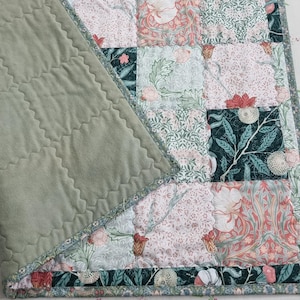 May include: A patchwork quilt with a sage green backing and a floral patterned front. The front features squares of fabric with various floral designs in shades of pink, green, and coral. The quilt is edged with a patterned border.