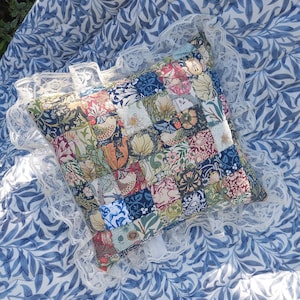 May include: A square patchwork cushion with a white lace trim. The cushion displays a variety of floral and bird patterns in shades of blue, red, green, and cream. It rests on a blue and white floral patterned fabric.