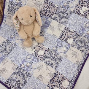 May include: A plush, tan bunny with floral-patterned ears rests on a patchwork quilt. The quilt features a mix of floral and geometric patterns in shades of blue, white, and gray, with a purple border.