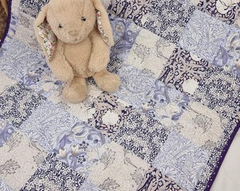 Könnte beinhalten: Ein beiges Stoffkaninchen liegt auf einer Patchwork-Decke. Die Decke zeigt florale und geometrische Muster in Blau-, Weiß- und Grautönen mit einem violetten Rand.