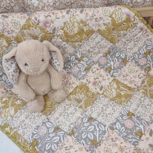 May include: A plush beige bunny sits on a patchwork quilt. The quilt features a floral pattern in shades of pink, gray, and gold. The bunny has floral patterned ears.
