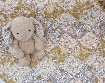 Könnte beinhalten: Ein beiges Plüschkaninchen sitzt auf einer Patchwork-Decke. Die Decke hat ein Blumenmuster in Rosa, Grau und Gold. Das Kaninchen hat Ohren mit Blumenmuster.