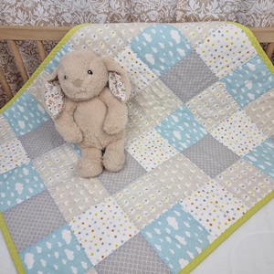 May include: A beige plush rabbit toy with floral-patterned inner ears sits on a patchwork baby quilt. The quilt features alternating squares of light blue with white clouds, grey with a subtle pattern, and white with multi-colored dots, all bordered by a bright lime green trim.