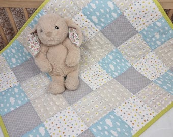 Könnte beinhalten: Ein beiges Plüschhasenspielzeug mit floral gemusterten Innenohren sitzt auf einer Patchwork-Babydecke. Die Decke besteht aus abwechselnden Quadraten in Hellblau mit weißen Wolken, Grau mit dezentem Muster und Weiß mit bunten Punkten, eingefasst von einer leuchtend lindgrünen Bordüre.