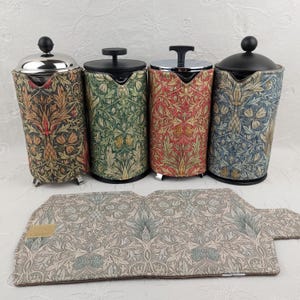 May include: Four French press coffee makers with patterned fabric sleeves in green, red, and blue. The fabric has a floral design. A matching fabric mat is in the foreground.