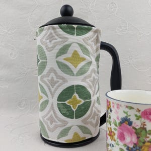 May include: A black French press coffee maker with a patterned cozy featuring green, yellow, and gray geometric designs. A floral patterned mug is next to the coffee maker. The background is a white, textured surface.