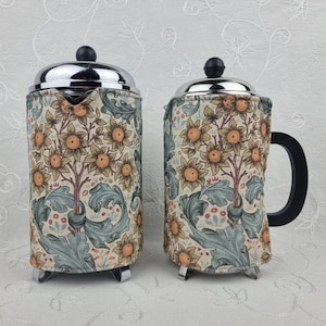 May include: Two French presses with a floral design. The presses have a stainless steel top and base, with a black handle on one. The fabric covering features a pattern of sunflowers, leaves, and branches in shades of yellow, green, and beige.
