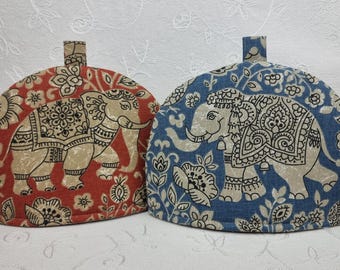 Könnte beinhalten: Zwei Teewärmer mit Elefantenmotiven. Einer ist rot mit einem beigen Elefanten und floralem Muster, der andere ist blau mit einem beigen Elefanten und floralem Muster. Beide haben oben eine Schlaufe für eine einfache Handhabung. Der Hintergrund ist weiß.