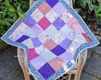 Könnte beinhalten: Eine quadratische Patchwork-Decke aus verschiedenen Stoffquadraten in Lila-, Rosa- und Weißtönen. Die Decke zeigt Muster mit kleinen Blumen und Zeichentricktieren und ist mit einer blaugrünen Bordüre mit rosa Blumenmuster eingefasst.