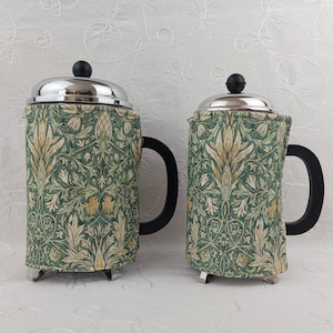 May include: Two French presses with stainless steel lids and black handles. The presses are covered in a green, cream, and yellow floral fabric. The presses are on a white, textured surface.