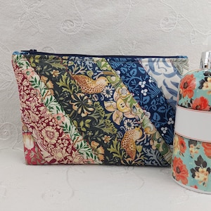 Peut inclure: Une trousse de maquillage matelassée avec fermeture éclair, présentant un motif patchwork de motifs floraux et d'oiseaux dans des tons de rouge, vert, bleu et crème. Une bouteille à motifs floraux assortie est à côté du sac.