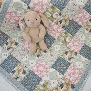 May include: A beige plush rabbit toy with long ears sits on a patchwork quilt. The quilt features a repeating pattern of square fabric patches in shades of dusty pink, sage green, and teal blue, decorated with floral and botanical leaf prints.