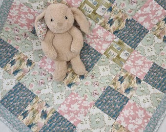 Könnte beinhalten: Ein beiges Plüschhasenspielzeug mit langen Ohren sitzt auf einer Patchwork-Decke. Die Decke besteht aus einem sich wiederholenden Muster aus quadratischen Stoffstücken in Altrosa, Salbeigrün und Blaugrün, verziert mit Blumen- und Blattmotiven.