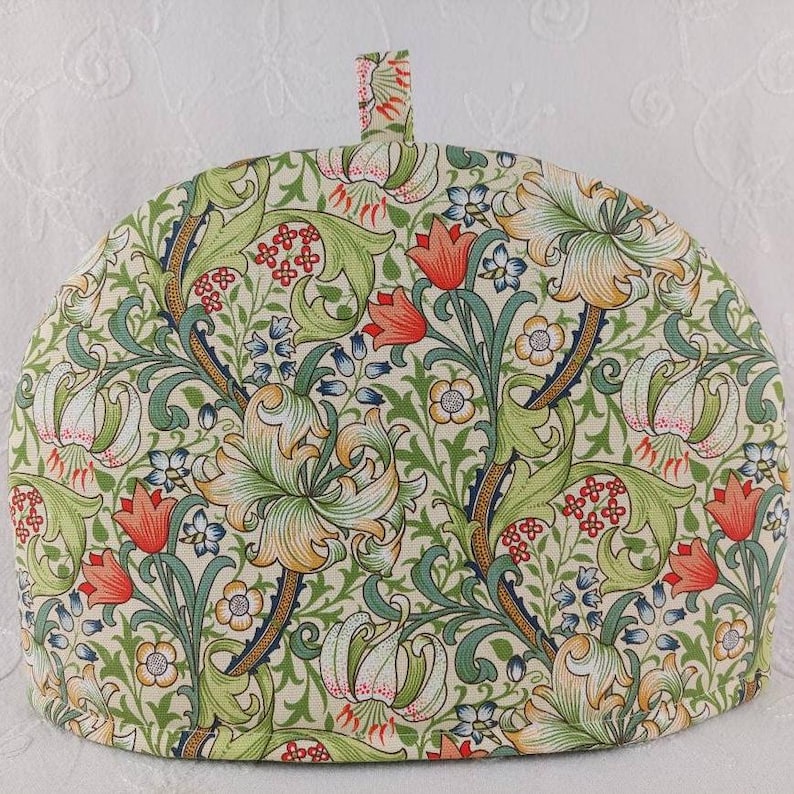 K&ouml;nnte beinhalten: Ein cremefarbener Teekoscher mit einem floralen Muster aus gr&uuml;nen Bl&auml;ttern, roten und orangen Blumen und wei&szlig;en Lilien. Das Muster erinnert an ein Vintage-Design.