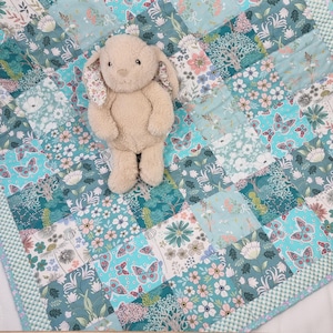 May include: A beige plush rabbit with floppy ears sits on a patchwork quilt. The quilt features a repeating pattern of teal, light blue, and white squares decorated with various floral motifs, green foliage, and butterflies in shades of pink and blue.