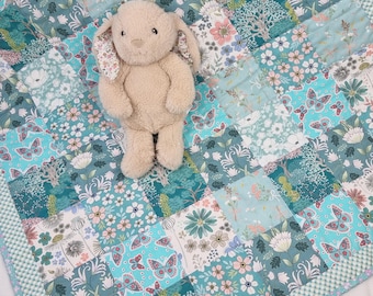 Könnte beinhalten: Ein beigefarbener Plüschhase mit Schlappohren sitzt auf einer Patchwork-Decke. Die Decke besteht aus einem sich wiederholenden Muster aus blaugrünen, hellblauen und weißen Quadraten, die mit verschiedenen Blumenmotiven, grünem Blattwerk und Schmetterlingen verziert sind.