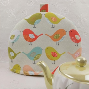 May include: A beige tea cosy with a colourful bird pattern. The birds are red, orange, yellow, green, and blue. The cosy has a loop handle.