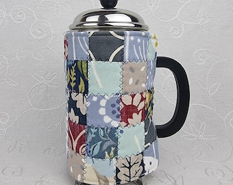 Könnte beinhalten: Eine French-Press-Kaffeebereiter mit einem Edelstahldeckel, einem schwarzen Knopf und einem schwarzen Griff. Die Glaskaraffe ist von einer Stoffhülle aus gesteppten, mehrfarbigen Patchwork-Quadraten in Blau-, Rot- und Cremetönen umgeben.