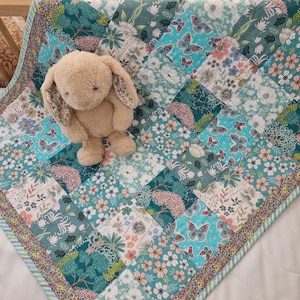May include: A patchwork quilt with a floral and butterfly pattern in shades of teal, pink, and white. A beige stuffed bunny sits on top of the quilt. The quilt has a striped border.