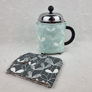 May include: A French press coffee maker with a light teal cozy featuring a white leaf pattern. A matching gray and white leaf patterned coaster is in the foreground. The coffee maker has a black handle and a silver lid.