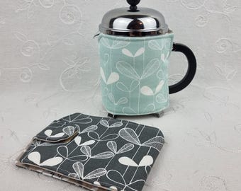 Könnte beinhalten: Eine French-Press-Kaffeemaschine mit einer helltürkisen Hülle mit weißem Blättermuster. Ein passender Untersetzer mit grauem und weißem Blättermuster befindet sich im Vordergrund. Die Kaffeemaschine hat einen schwarzen Griff und einen silbernen Deckel.