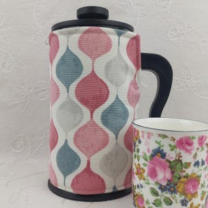 May include: A French press coffee maker with a black handle and lid, and a patterned cosy in pink, grey, and white. A floral-patterned mug is next to the coffee maker. The cosy has a repeating teardrop design.