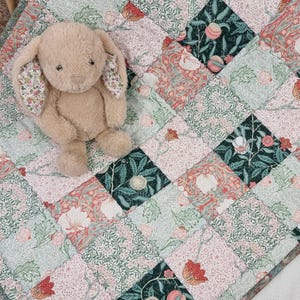 May include: A plush, tan bunny toy with patterned ears rests on a patchwork quilt. The quilt features a floral design in shades of pink, green, and red. The quilt is made of square patches.