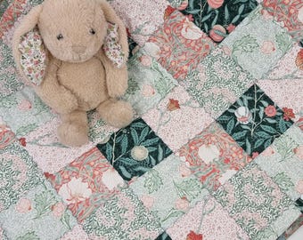 Könnte beinhalten: Ein kuscheliges, hellbraunes Spielzeugkaninchen mit gemusterten Ohren liegt auf einer Patchwork-Decke. Die Decke hat ein Blumendesign in Rosa, Grün und Rot. Die Decke besteht aus quadratischen Flicken.