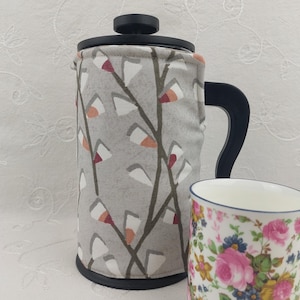 May include: A French press coffee maker with a gray fabric cozy featuring a white, orange, and red tree branch pattern. A floral patterned teacup is next to the coffee maker. The coffee maker has a black handle and lid.