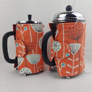 May include: Two orange and white patterned fabric coffee press cozies. The cozies are made of a soft, quilted fabric with a floral design. The cozies are designed to fit standard-sized coffee presses.