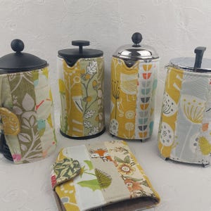 May include: Four French press coffee makers with quilted fabric sleeves. The sleeves feature a patchwork design with floral, bird, and leaf patterns in yellow, green, and white. A matching quilted cozy is also present.