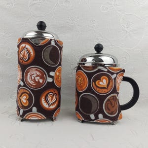 May include: Two coffee press covers with a brown background and a pattern of coffee art in shades of brown, orange, and white. The covers have a stainless steel top and a black handle. One is tall and cylindrical, the other is shorter and wider.