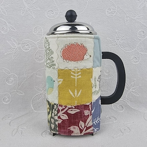 May include: A French press coffee maker with a patchwork fabric cozy. The cozy features a variety of patterned squares with images of a bird, hedgehog, and botanical designs. The handle is black, and the lid is silver.