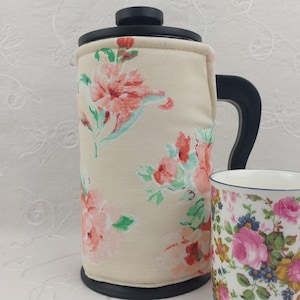 May include: A French press coffee maker with a floral insulated cozy and a matching floral mug. The cozy is beige with pink and red flowers and green leaves. The mug has a similar floral pattern with pink roses and blue accents.