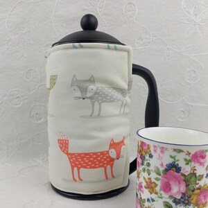 May include: A cream-colored French press with a black handle and lid, featuring a fabric cozy with fox illustrations in orange and gray. A floral-patterned mug is next to the press.
