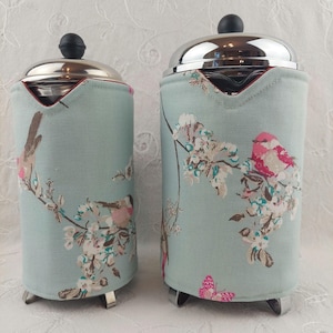 May include: Two French press coffee makers with a light blue fabric cover with a floral pattern featuring birds and butterflies.