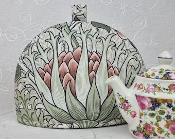 Könnte beinhalten: Ein floraler Teewärmer und eine passende Teekanne. Der Teewärmer hat ein botanisches Design in Grün-, Rosa- und Beigetönen. Die Teekanne ist weiß mit rosa und blauen Blumenmustern. Der Teewärmer hat einen kleinen Griff oben.