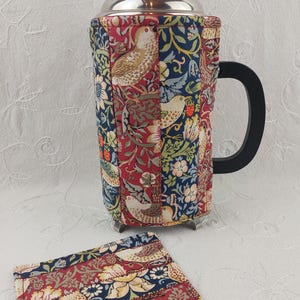 May include: A red, blue, and green fabric coffee press cozy with a bird and floral pattern. The cozy is on a white surface.