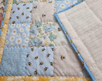 Könnte beinhalten: Eine gesteppte Babydecke mit einem Patchwork-Design aus hellblauen, weißen und beigen Quadraten. Die Stoffmuster zeigen Hummeln, Gänseblümchen und florale Motive. Die Decke ist mit gelbem Blumenstoff eingefasst und hat eine weiße Sherpa-Rückseite.