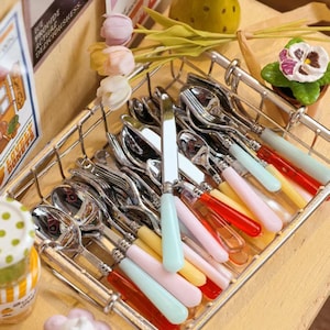 May include: A rectangular wire basket filled with assorted stainless steel cutlery featuring colorful translucent acrylic handles in shades of pastel pink, light blue, yellow, and red, arranged neatly alongside decorative artificial flowers.