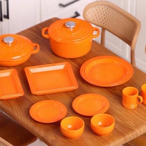 Könnte beinhalten: Ein Set orangefarbener Miniatur-Küchenutensilien, bestehend aus Töpfen, Tellern, Schalen und Tassen. Das Set ist auf einem Holztisch angeordnet.