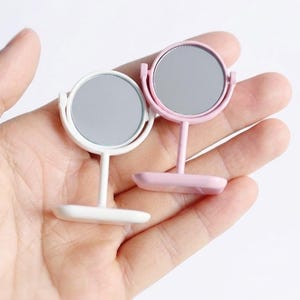 May include: Two miniature vanity mirrors, one white and one pink, are held in a hand. Each mirror has a round, silver-colored reflective surface and a stand. The mirrors are small, likely for a dollhouse or craft project.