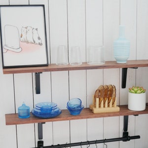 May include: Two wooden shelves with black metal brackets, holding various items including a framed illustration of three cats watching a television, two clear glass cups, a blue and white striped vase, a blue glass jar, a set of blue glass plates, a blue glass bowl, a wooden coaster with six brown glass coasters, a small white planter with a green succulent, and a larger white planter with a green succulent.